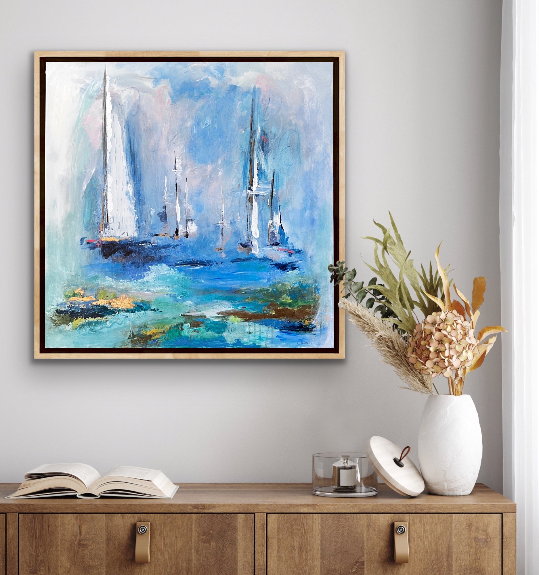 Blue, cream and gold coastal abstract mixed media painting, 20" x 20" framed in natural wood float frame. Mixed media painting with acrylic paint, gold leaf, pastel, charcoal and vintage piano roll paper collaged into the sail of an abstract sailboat. Calming coastal painting against white wall.