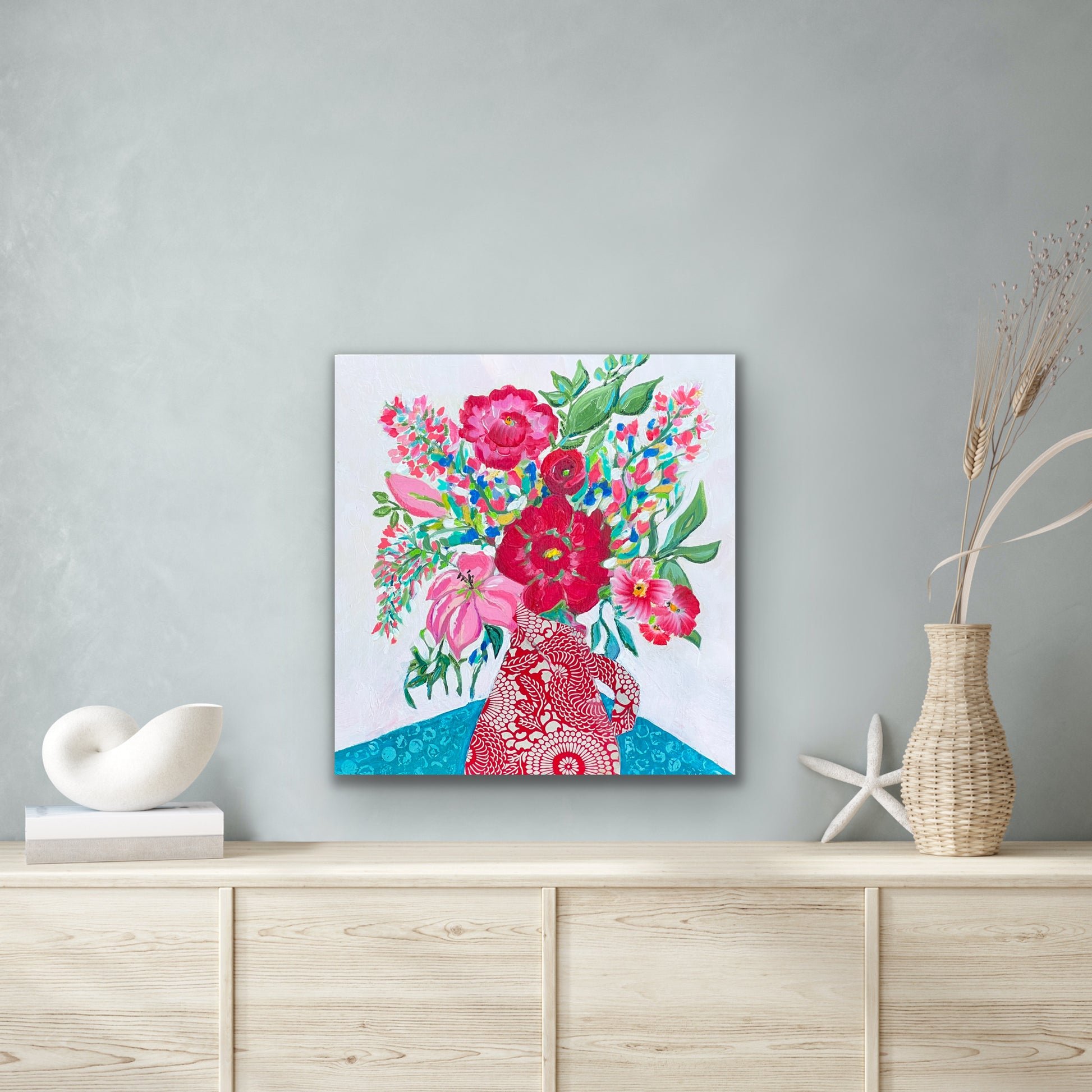 This is a 12" x 12" abstract floral painting. The main colors are red, pink and turquoise, with some green leaf accents throughout. The painting is a festive, loose bouquet of flowers spilling out of a pitcher, sitting on a turquoise tablecloth with an abstract pattern on it. The pitcher is cut from Japanese origami paper in a red and white floral motif.  The painting is shown against a light gray wall with some coastal style room accents.