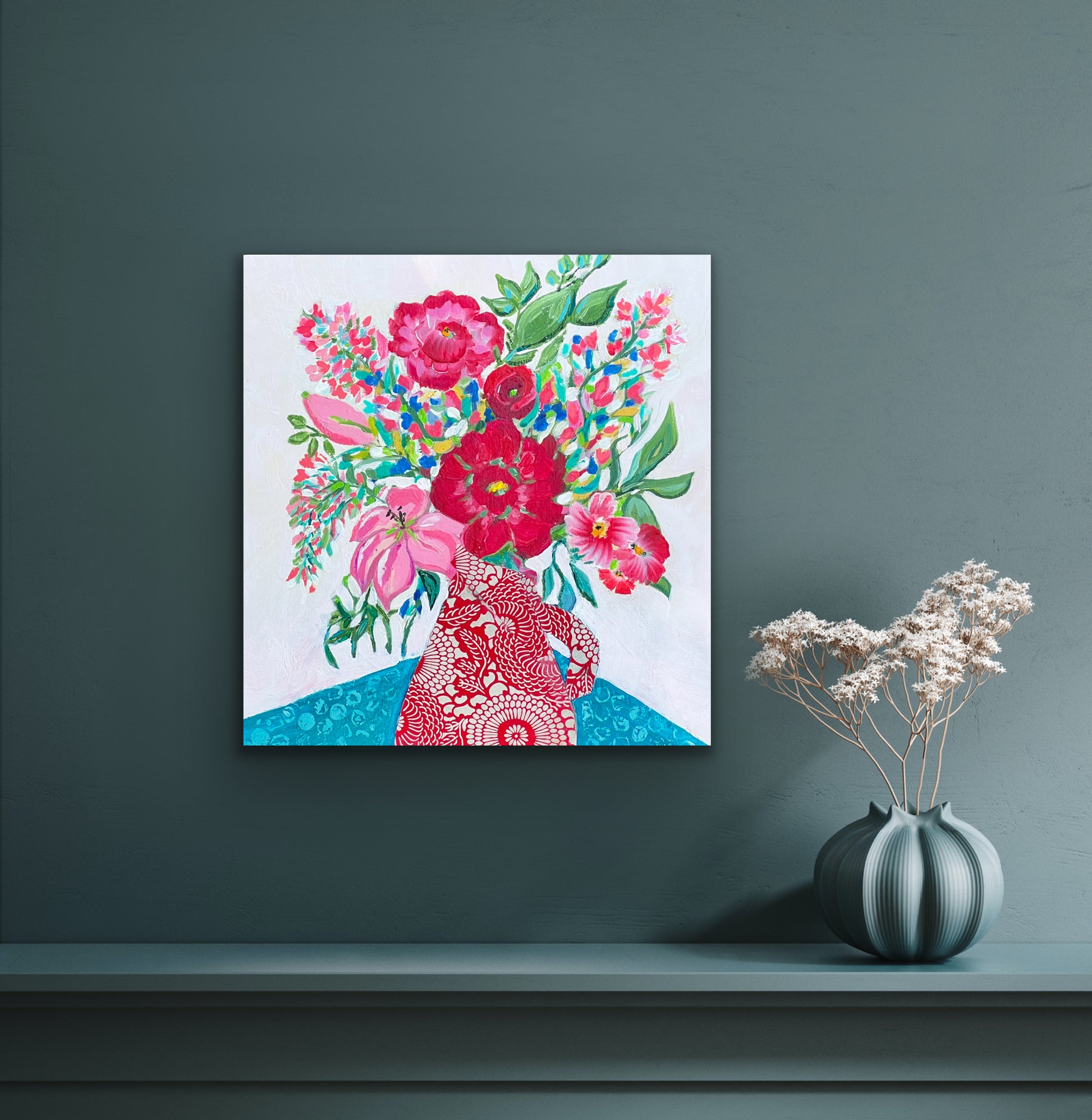 This is a 12" x 12" abstract floral painting. The main colors are red, pink and turquoise, with some green leaf accents throughout. The painting is a festive, loose bouquet of flowers spilling out of a pitcher, sitting on a turquoise tablecloth with an abstract pattern on it. The pitcher is cut from Japanese origami paper in a red and white floral motif. The painting is shown against a gray-green wall. 